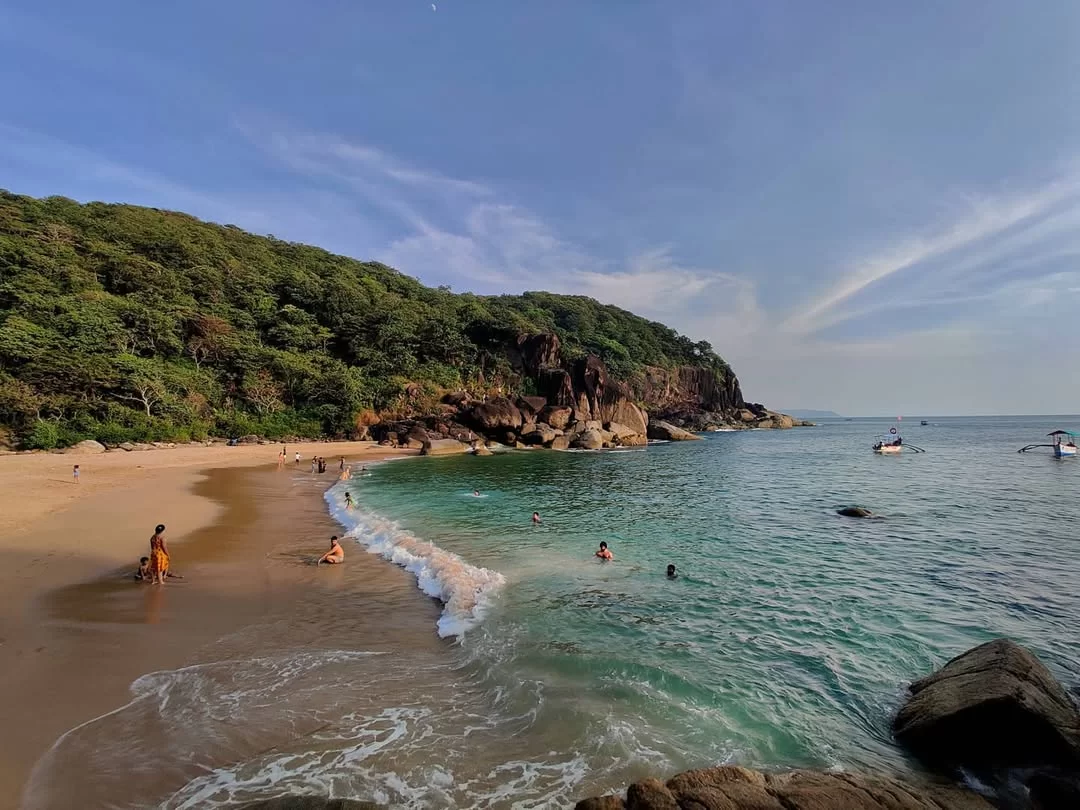 Vibrant turquoise waters Butterfly Beach South Andaman sunny day, featuring rocky cliffs lush green hills people swimming boats sandy shore, perfect Andaman Islands beach tour package. 