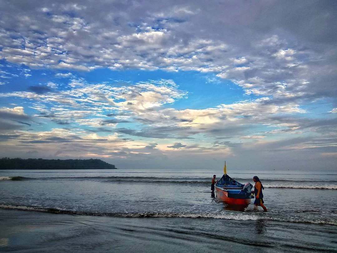 Fishermen pulling orange blue boat ashore at Caranzalem Beach Goa during cloudy skies, featuring waves palms, perfect adventure experience Goa beach tour package.