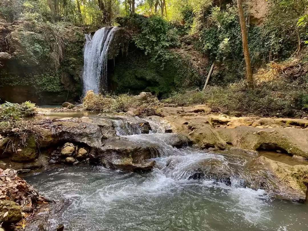 Jim Corbett Falls scenic waterfall terraced rocks green pool ivy cliffs, rippling cascade limestone boulders vines trees golden light, perfect Uttarakhand tour packages.