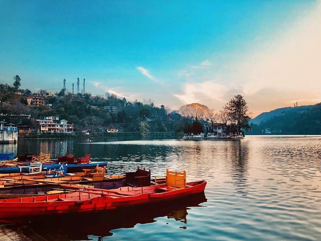 Bhimtal Lake sunset foreground with colorful rowboats reflecting on water, distant hills resorts & chimneys under orange sky, perfect romantic experience Uttarakhand tour packages.