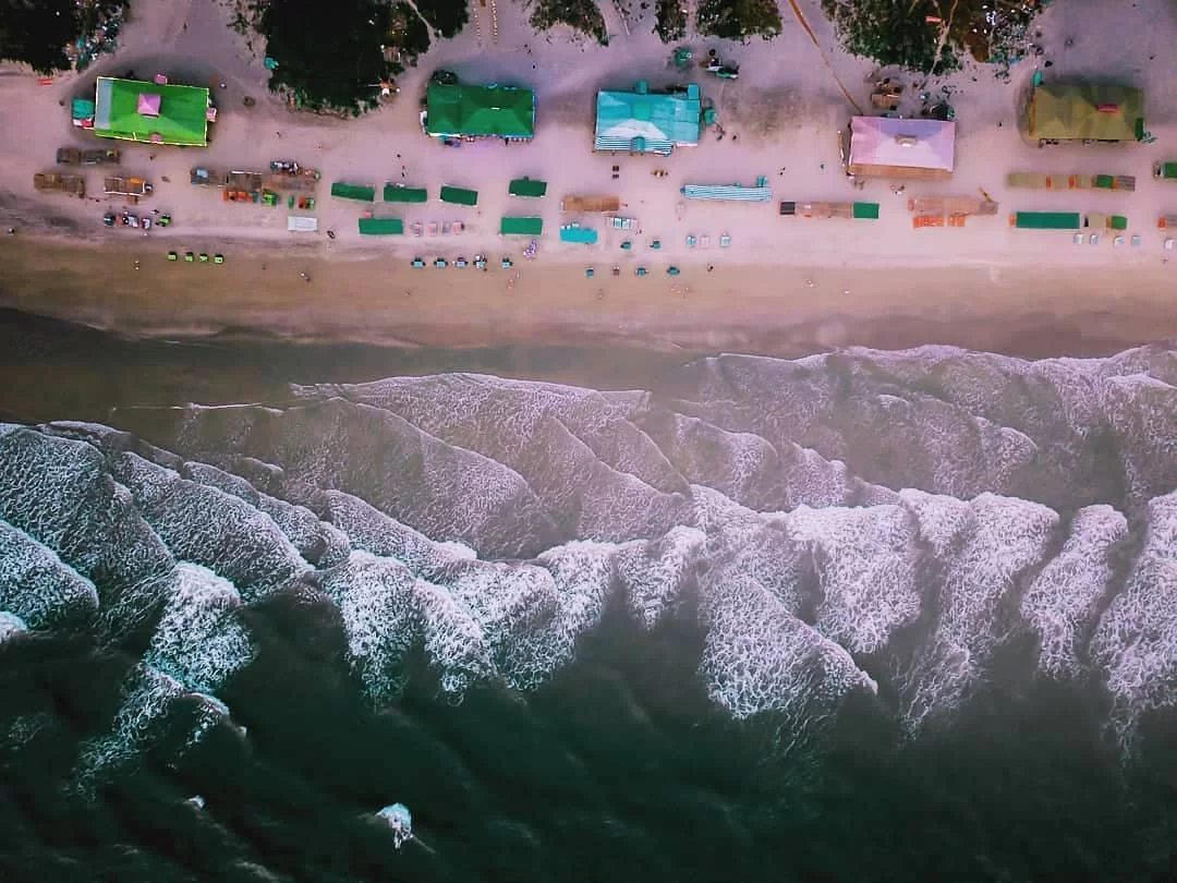 Drone aerial view of Mandrem Beach North Goa during golden hour sunset, featuring colorful shacks beach umbrellas chairs palm trees white sandy shore Arabian Sea waves, perfect serene beach shack experience with Mandrem Beach Goa tour package.
