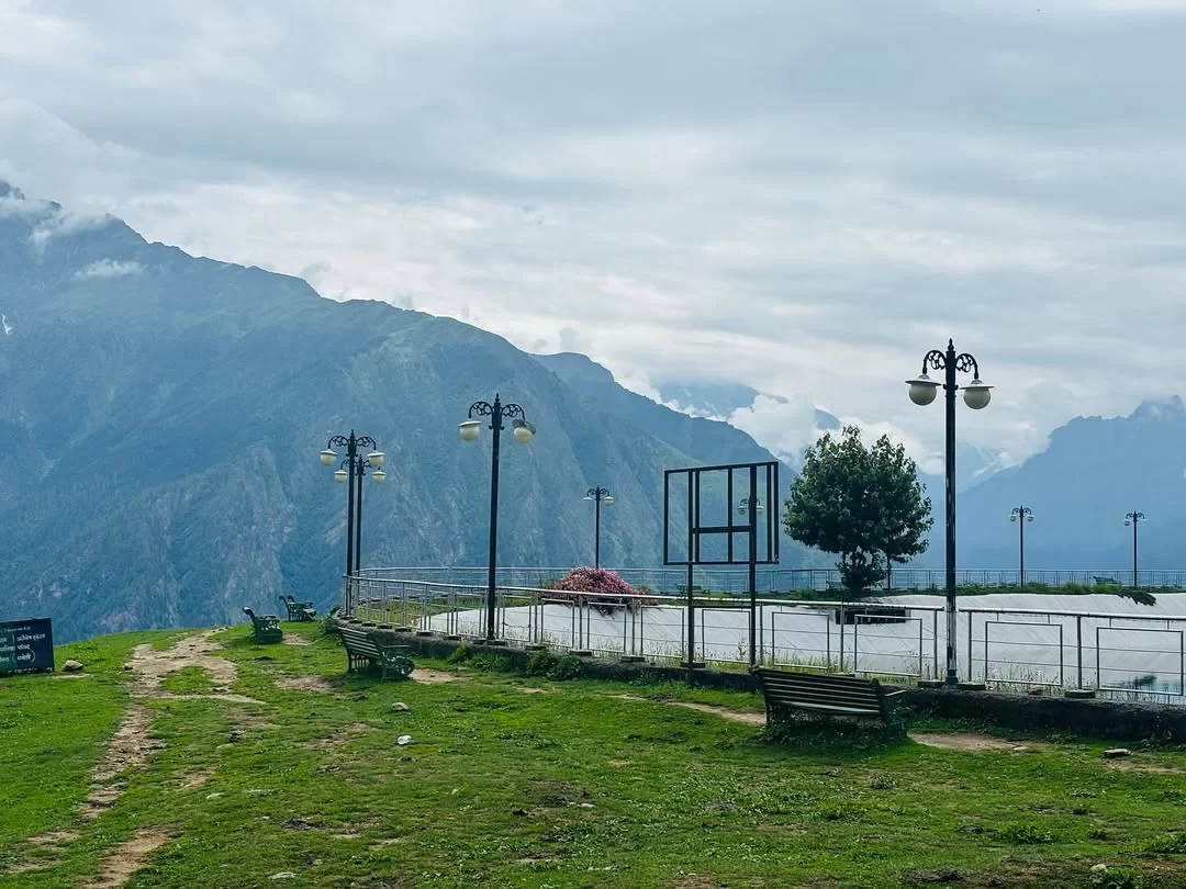 Auli Artificial Lake area Uttarakhand cloudy day, featuring basketball court benches lamps tree railing misty Himalayan peaks backdrop, perfect adventure experience Uttarakhand tour packages.