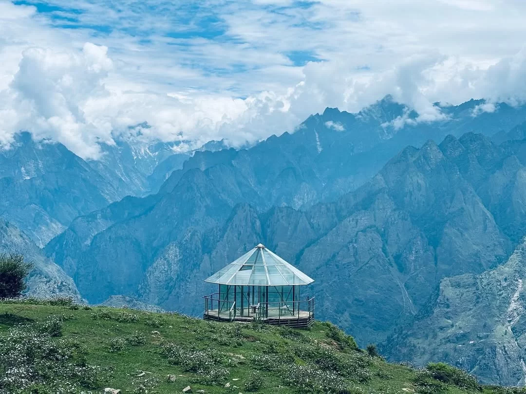 Auli viewpoint pavilion Uttarakhand cloudy skies, featuring glass gazebo overlooking green meadows snowcapped Himalayan peaks, perfect adventure experience Uttarakhand tour packages.