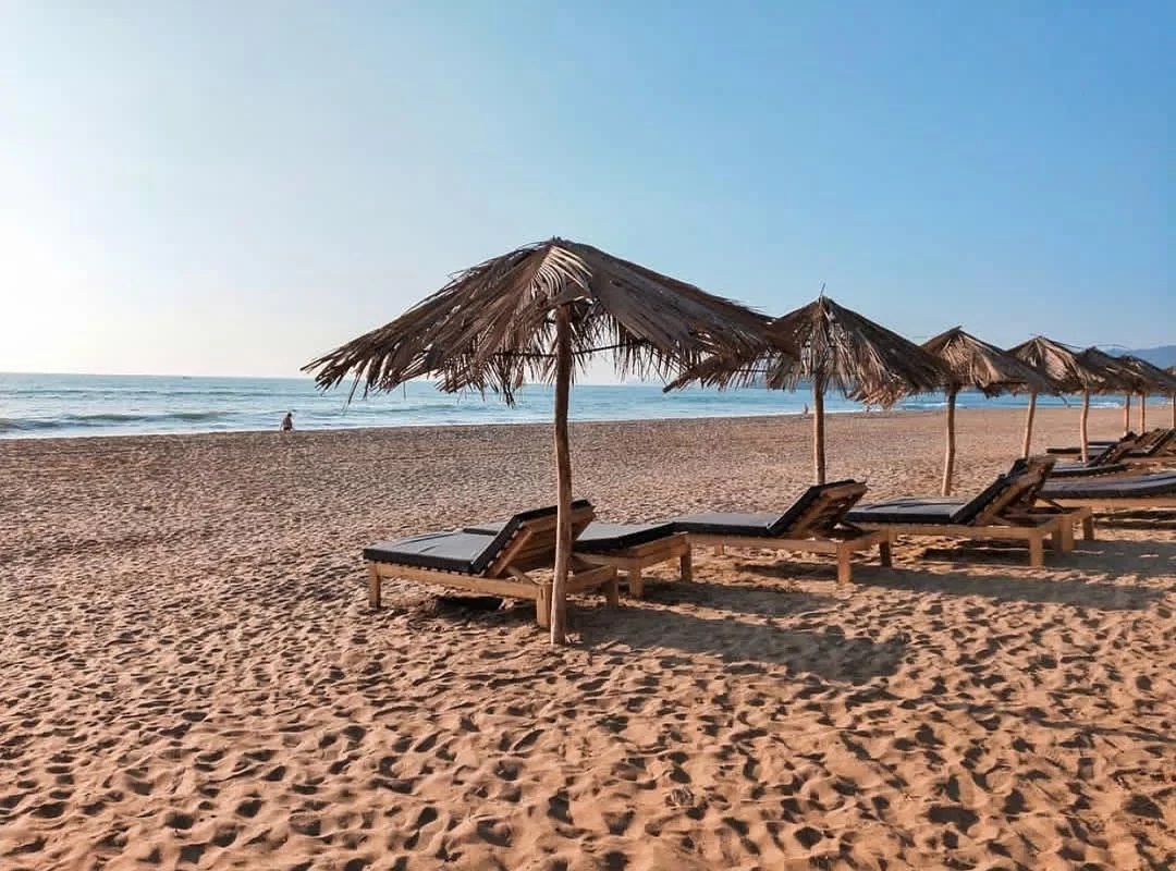 Row of palm umbrellas with sunbeds at Agonda Beach Goa during golden hour, featuring calm sea lone figure waves, perfect romantic Goa tour package. 
