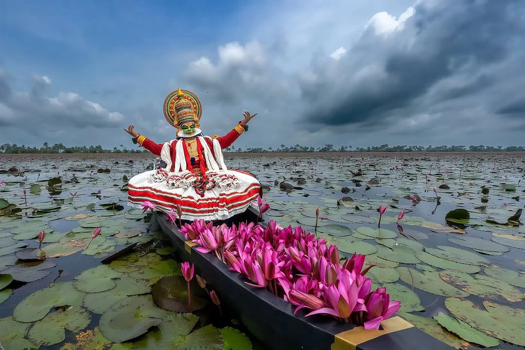 Kerala backwaters Kathakali performer arms raised elaborate green face paint golden headpiece white red skirt costume black boat pink lotus flowers green lily pads cloudy sky, traditional dance cultural tour package.