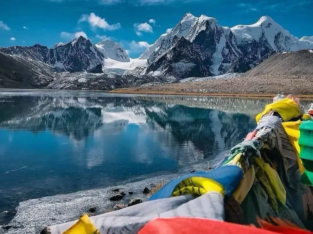 Tsomgo Lake prayer flags during sunny winter day, featuring colorful fluttering flags reflecting on turquoise lake with snowy glacier mountains backdrop, perfect adventure Sikkim tour packages.