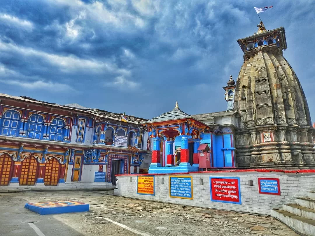 Ukhimath Omkareshwar Temple towering stone shikhara colorful blue red yellow wood carved facade cloudy skies flag fluttering, perfect Kedarnath winter Shiva temple, Uttarakhand tour packages.