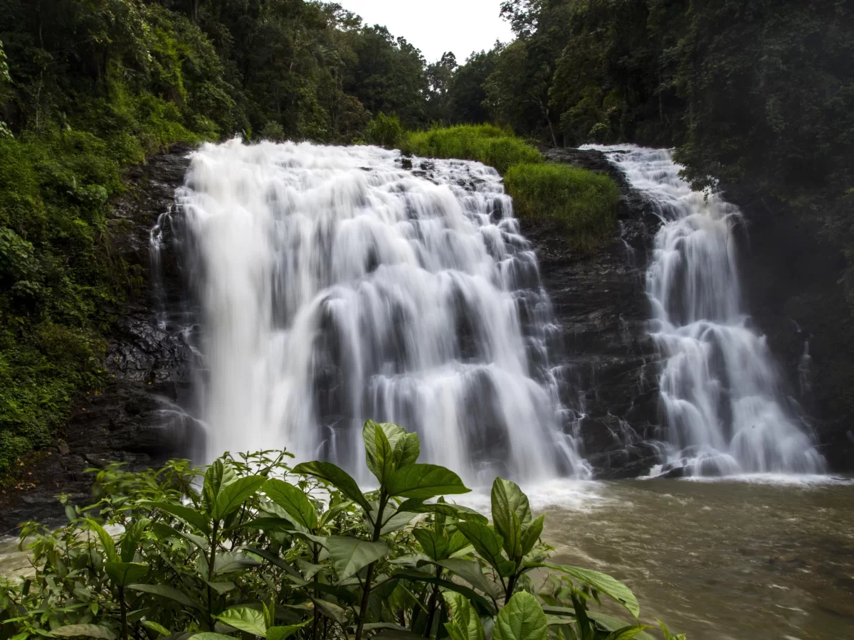 Abbey Falls at Coorg during cloudy day, featuring multi-tiered waterfall, lush forests, rocky cascade, river base, perfect nature experience Karnataka tour packages.