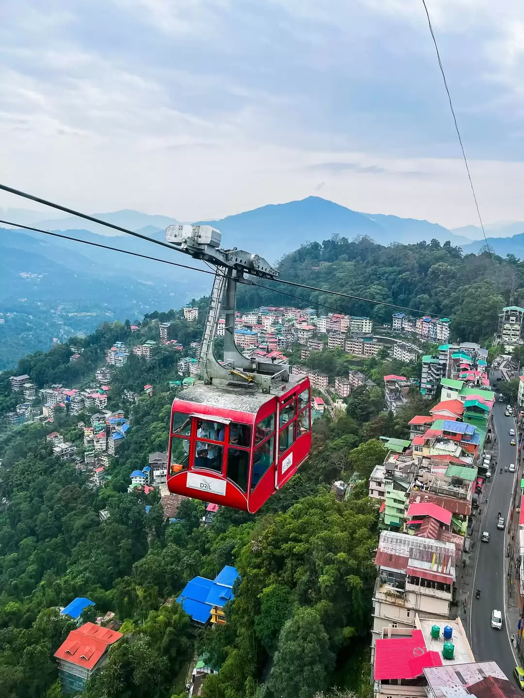 Red cable car gliding above colorful hillside cityscape in Gangtok with panoramic mountain views, an exciting attraction featured in Sikkim tour packages.