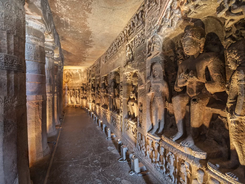 Ajanta Caves Maharashtra UNESCO rock-cut Buddhist caves interior hallway with ancient intricately carved stone pillars and Buddha sculptures illuminated by lamps, iconic heritage tour package.