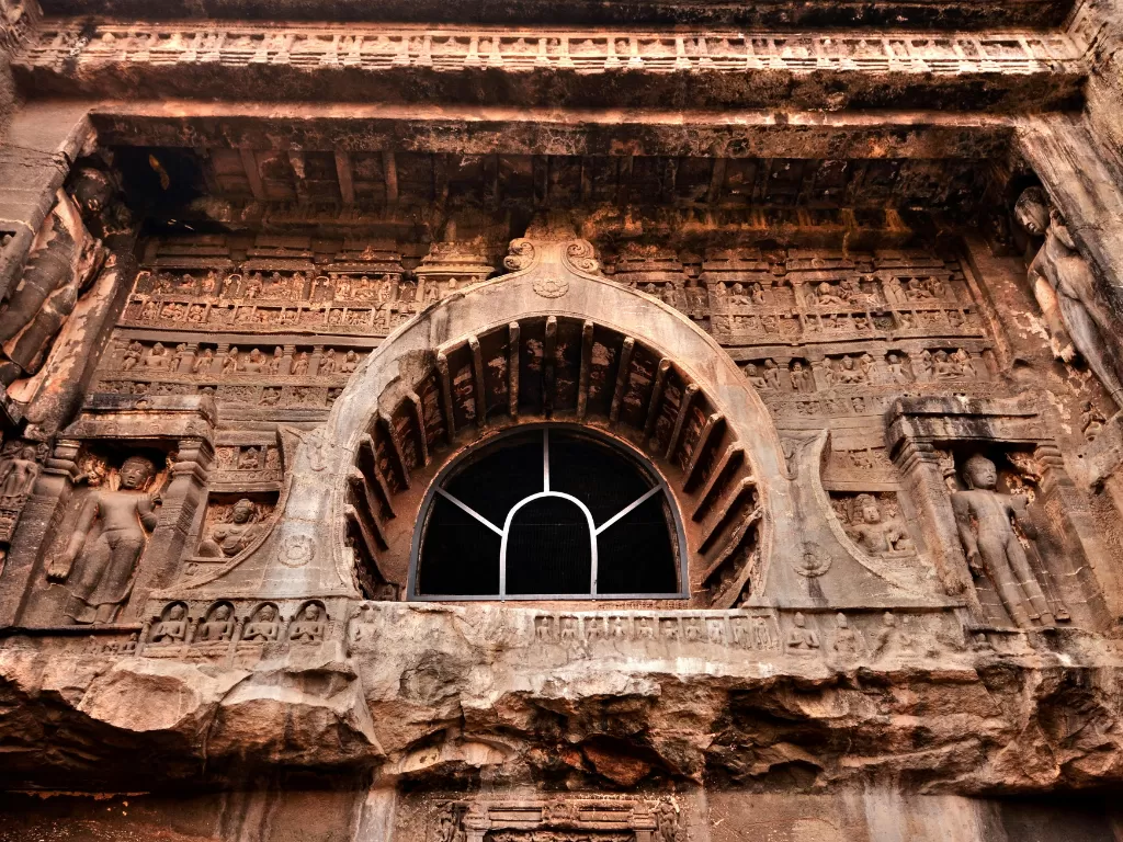 Ajanta Caves Maharashtra close-up ornate rock-cut chaitya arch facade with detailed Jain Tirthankara sculptures horseshoe window eroded stonework dramatic warm lighting, timeless UNESCO Buddhist-Jain art heritage tour package.