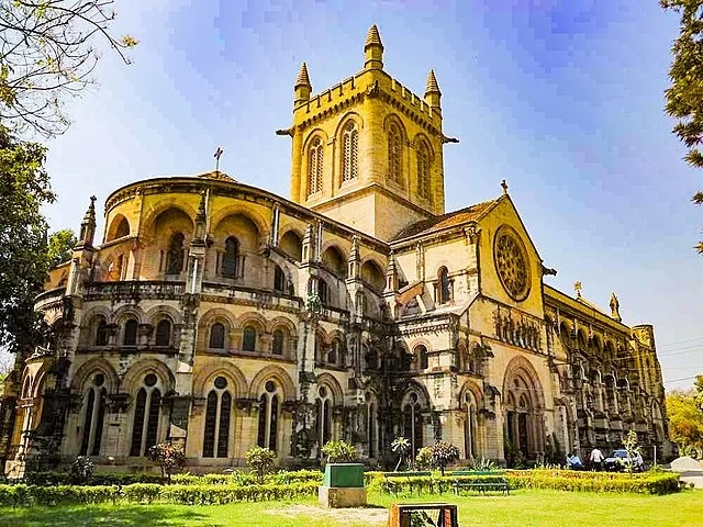 All Saints Cathedral sunny sky, Gothic yellow stone towers rose window arches trees lawns cars, perfect Uttar Pradesh tour package.