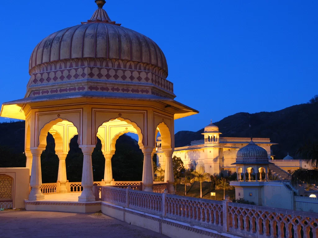 Illuminated sandstone pavilion with dome and arches at night overlooking palaces and hills at Amber Fort in Jaipur Rajasthan under twilight blue sky, perfect Rajasthan tour package.