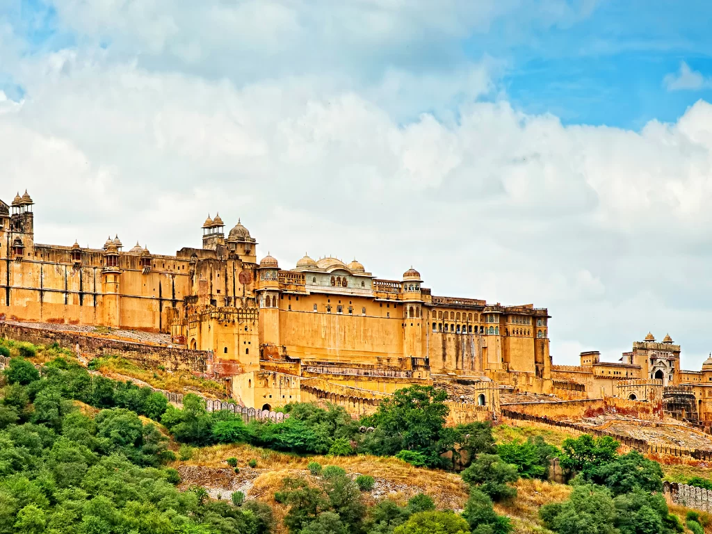Full facade view of Amber Fort (Amer Fort) in Jaipur Rajasthan on Aravalli hillside with multi-tiered yellow sandstone palaces, towers amid greenery under partly cloudy blue sky, perfect Rajasthan tour package.