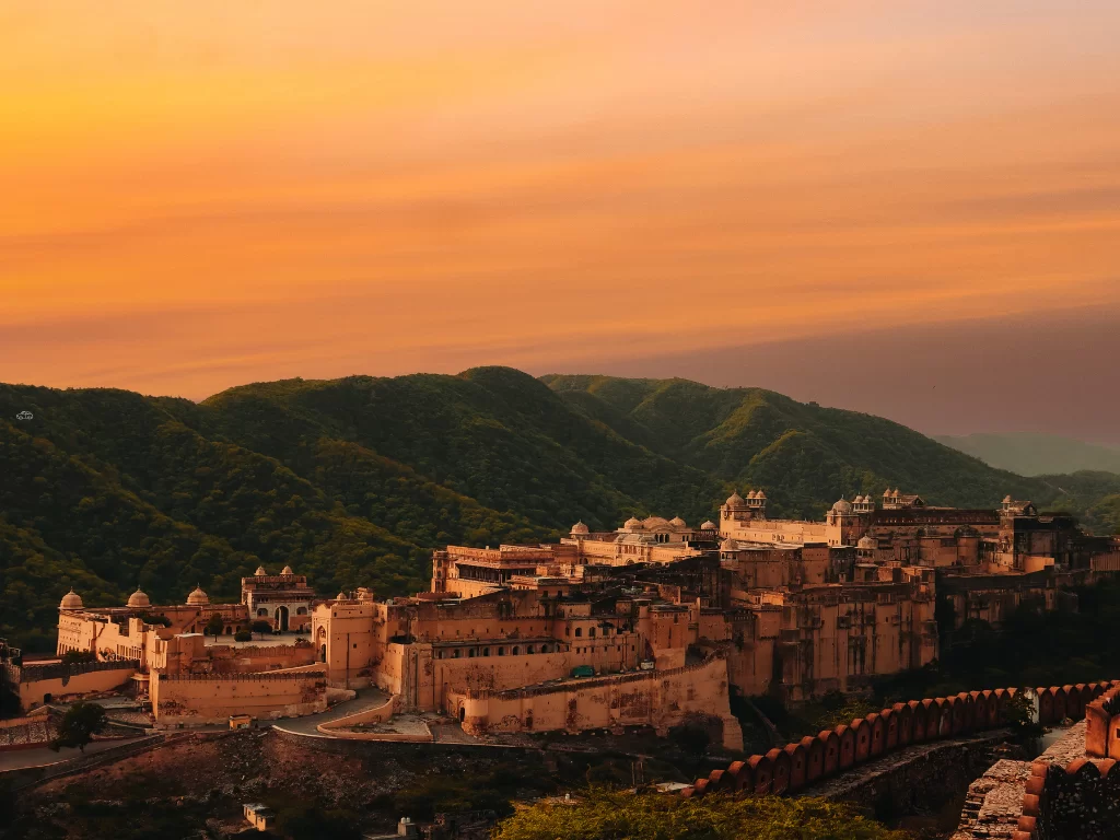 Sunset panoramic view of Nahargarh Fort in Jaipur Rajasthan atop Aravalli hills with pinkish sandstone structures, walls amid green slopes under orange purple sky, perfect Rajasthan tour package