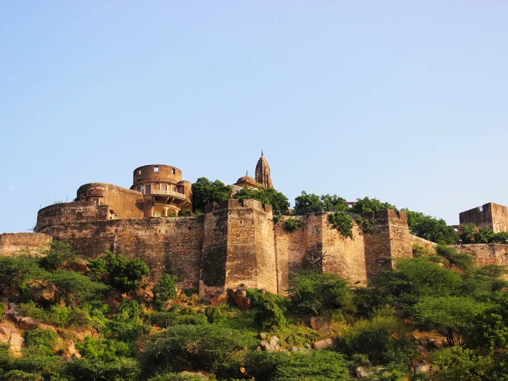 Panoramic view of Jaigarh Fort in Jaipur Rajasthan with round bastions, tall temple spire, crenellated walls amid lush green hill under clear blue sky, perfect Rajasthan tour package.