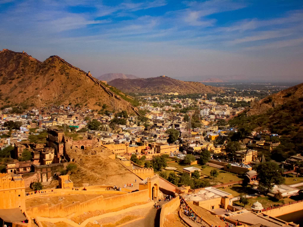 Panoramic aerial view of Amer Fort (Amber Fort) in Jaipur Rajasthan overlooking Maota Lake, colorful old town, Aravalli hills under golden sunset sky, perfect Rajasthan tour package.