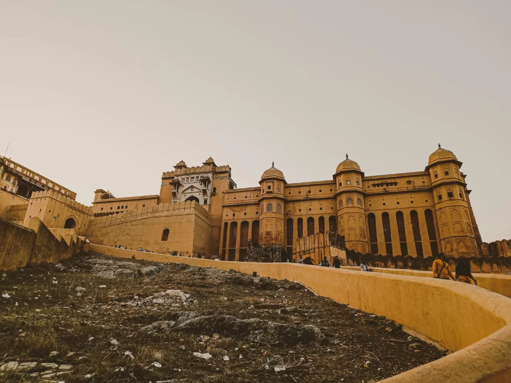 Golden hour view of Amer Fort (Amber Fort) in Jaipur Rajasthan showcasing majestic sandstone facade, domes, ramparts and hillside path with visitors under hazy sky, perfect Rajasthan tour package