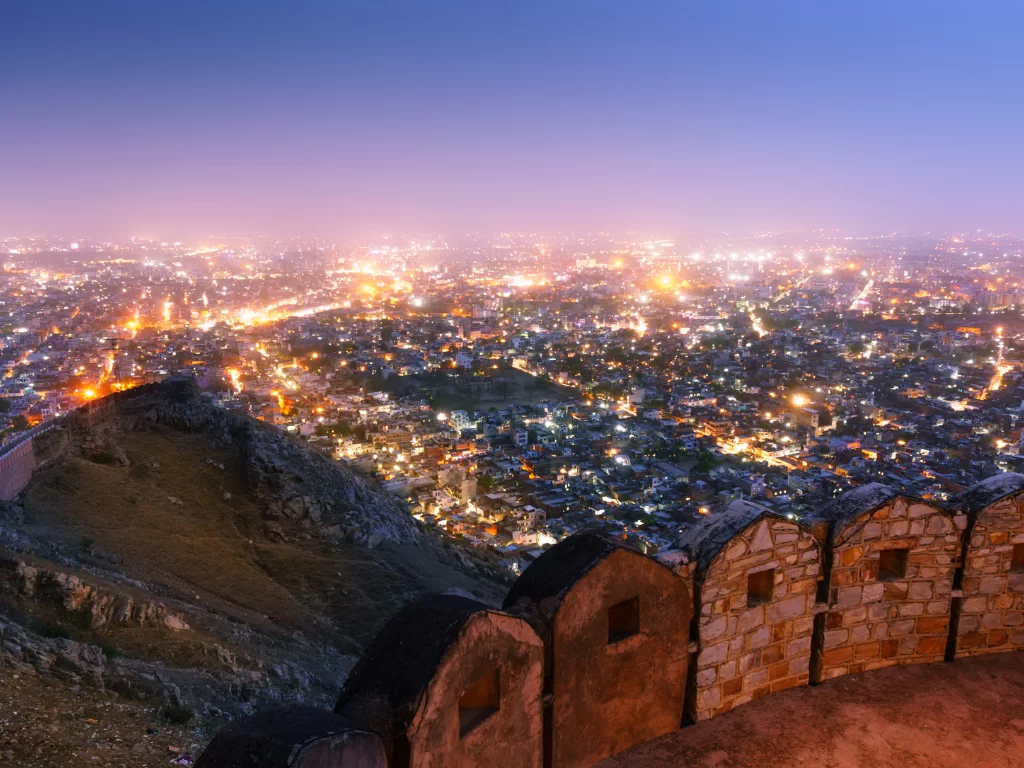Night panoramic view from Nahargarh Fort overlooking twinkling Jaipur city lights, Aravalli hills and fort walls under twilight purple sky, perfect Rajasthan tour package.