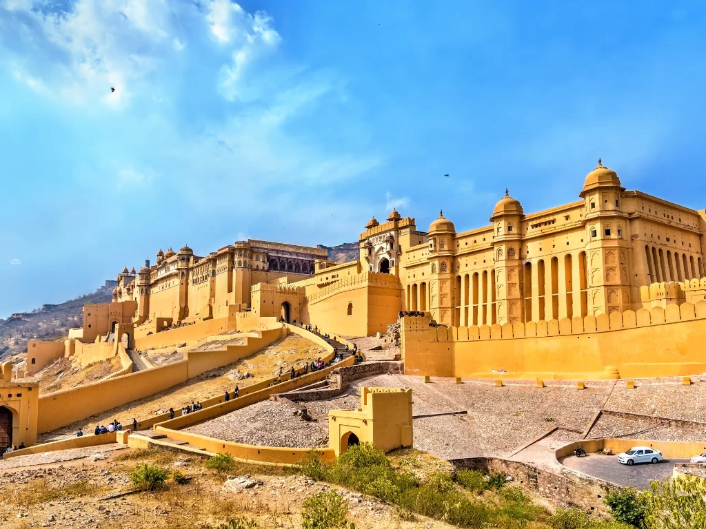 Daytime panoramic view of Amber Fort (Amer Fort) in Jaipur Rajasthan with expansive yellow sandstone walls, gates, towers on hillside amid hills and blue sky with clouds, perfect Rajasthan tour package.