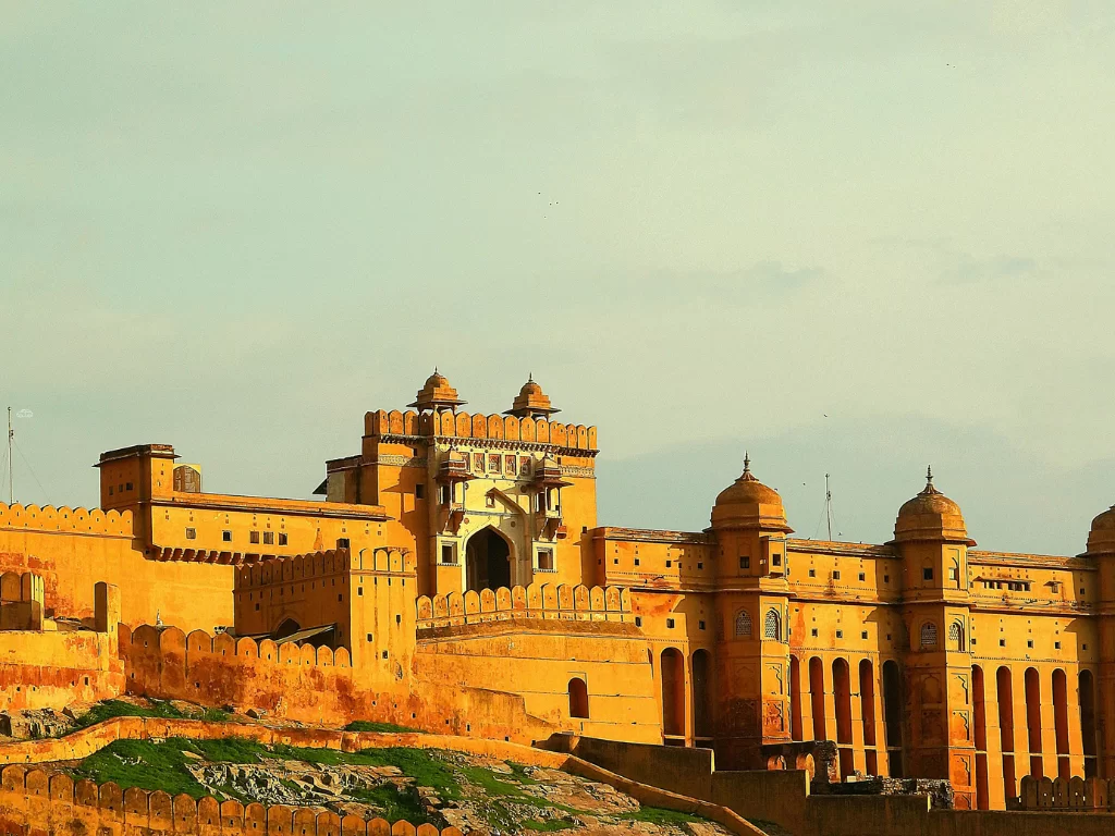 Golden hour view of Amber Fort main gate in Jaipur Rajasthan with towering yellow sandstone entrance, domes, walls on hill with greenery and hazy sky, perfect Rajasthan tour package.