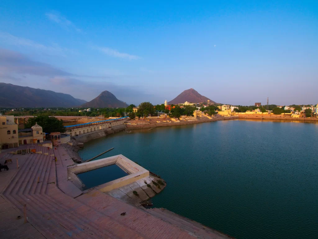 Ana Sagar Lake Ajmer Rajasthan at golden hour under blue skies with crescent moon, featuring ghats temples distant Aravalli hills and vibrant waters, perfect heritage Rajasthan tour package.