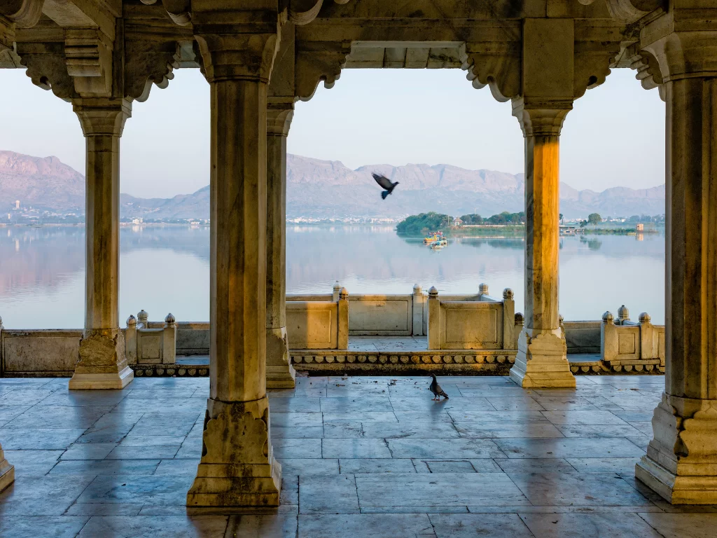 Jai Mandir pavilion in Jal Mahal Palace Jaipur Rajasthan at dawn with marble columns framing serene Man Sagar Lake distant Aravalli hills flying birds and misty reflections, iconic heritage Rajasthan tour package view