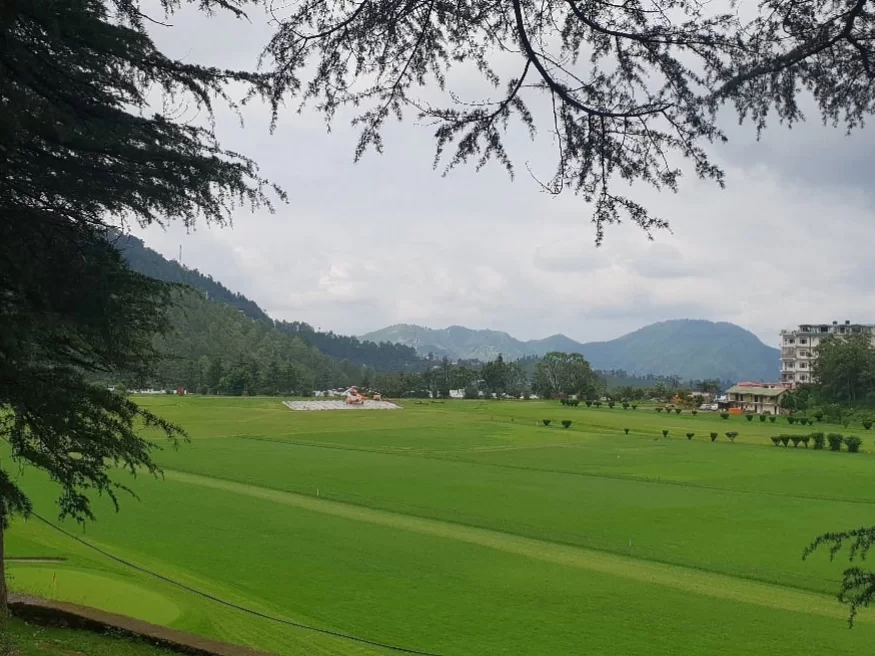 Annadale Ground in Shimla featuring a vast green meadow surrounded by pine trees and hills under a cloudy sky.