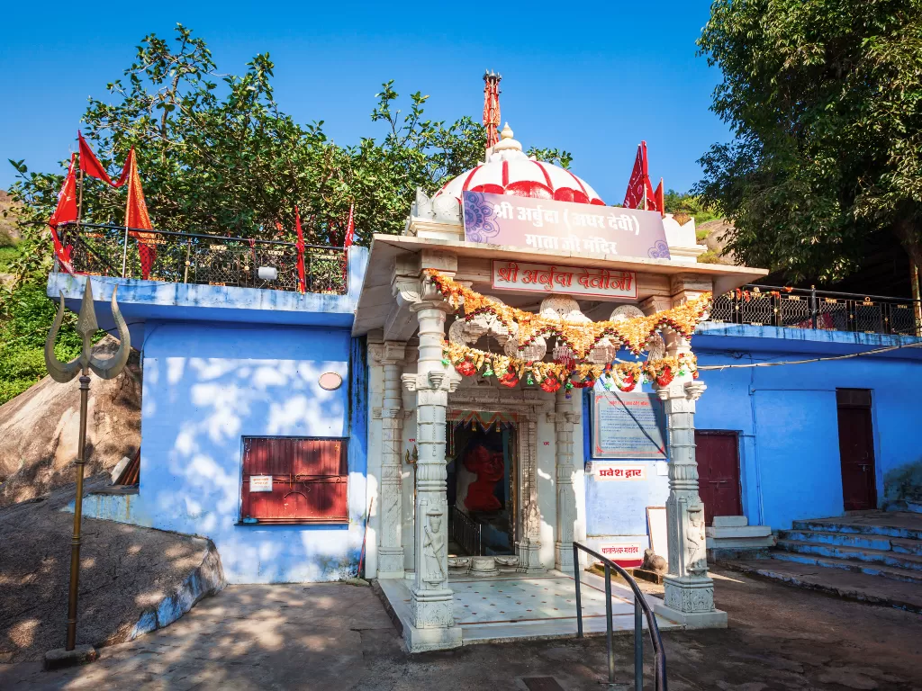 Arbuda Devi Temple blue facade dome flags marigold garlands Mount Abu, white shikhara trishul Hindi sign rocky hill trees sunny sky, perfect Rajasthan tour packages.