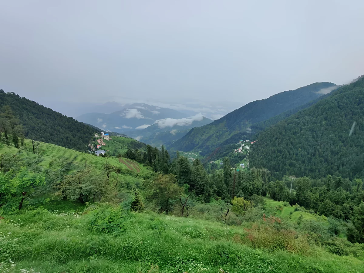 Bakrota Hills in Dalhousie featuring rolling green valleys, dense pine forests, scattered hillside houses, and mist-covered mountain ranges under a cloudy sky.