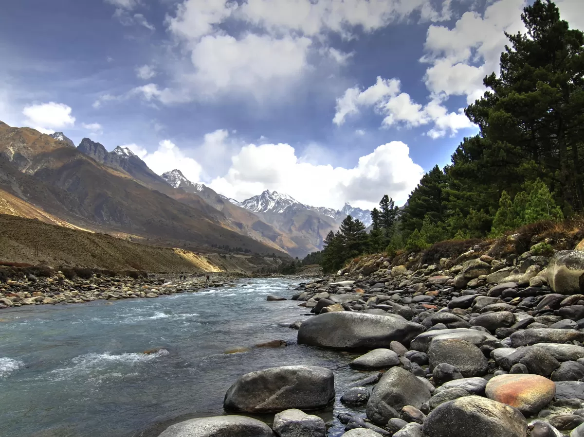 Baspa River Chitkul Village Kinnaur Himachal Pradesh during partly cloudy blue skies, featuring turquoise rapids large boulders pine forested snowy Himalayan peaks backdrop, perfect cultural experience Himachal tour packages.