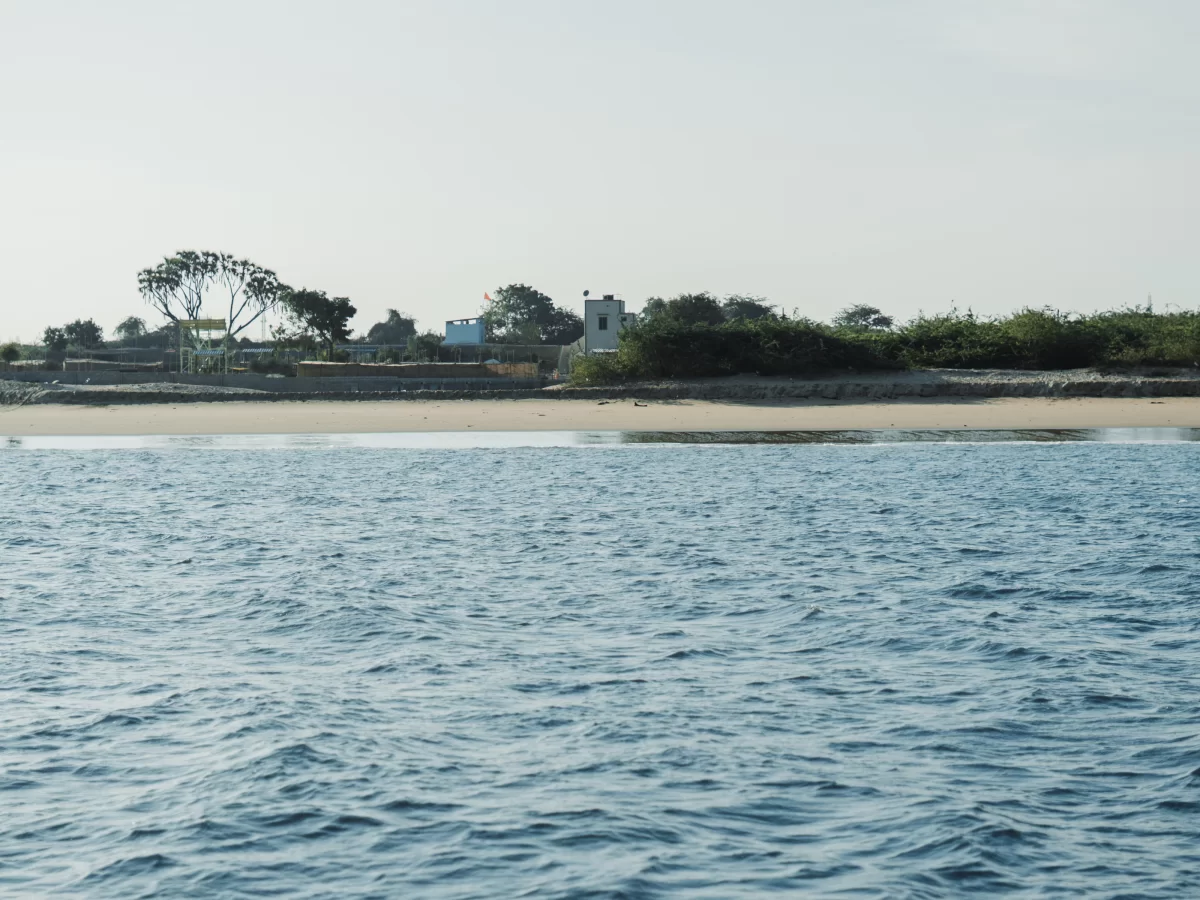 Beyt Dwarka island beach during clear day, featuring sandy shore with distant buildings, palm trees, and turquoise Arabian Sea, perfect pilgrimage experience with Gujarat tour packages.