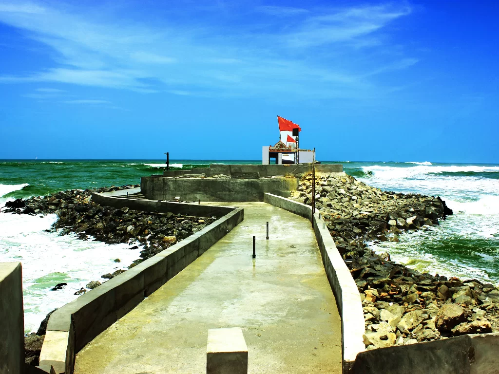 Bhadkeshwar Mahadev Temple Dwarka concrete pathway leading to white shrine with red saffron flags amid crashing Arabian Sea waves on rocky breakwater under clear blue skies, unique ocean Shiva temple perfect for Gujarat tour packages.