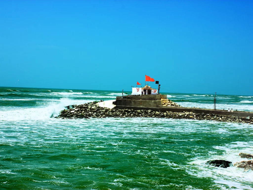 Bhadkeshwar Mahadev Temple Dwarka on rocky breakwater amid crashing Arabian Sea waves under clear blue skies with red saffron flags, unique sea Shiva shrine perfect for Gujarat tour packages.
