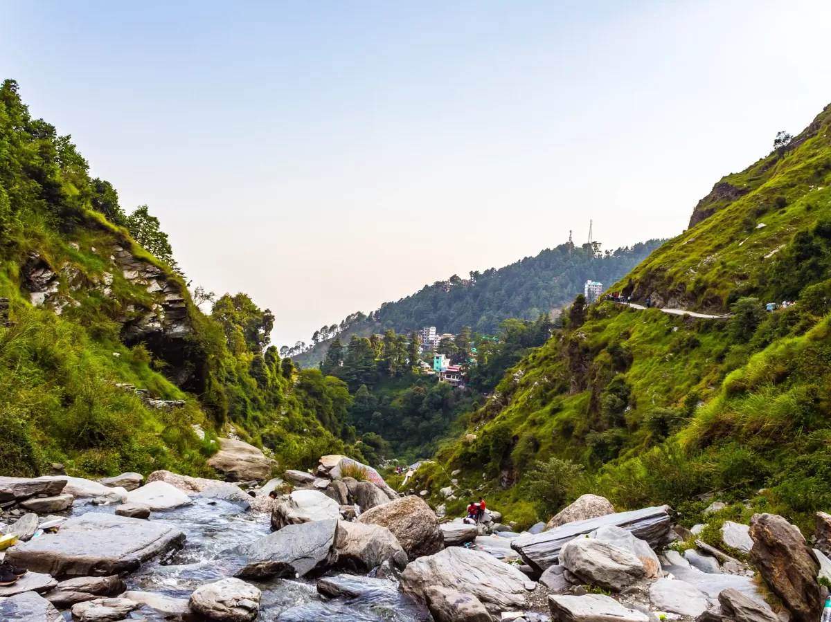 Trekkers on Bhagsu Nag Waterfall pathway at Dharamshala during golden hour, featuring lush green valleys, rocky stream, hills, perfect adventure Himachal Pradesh tour package.