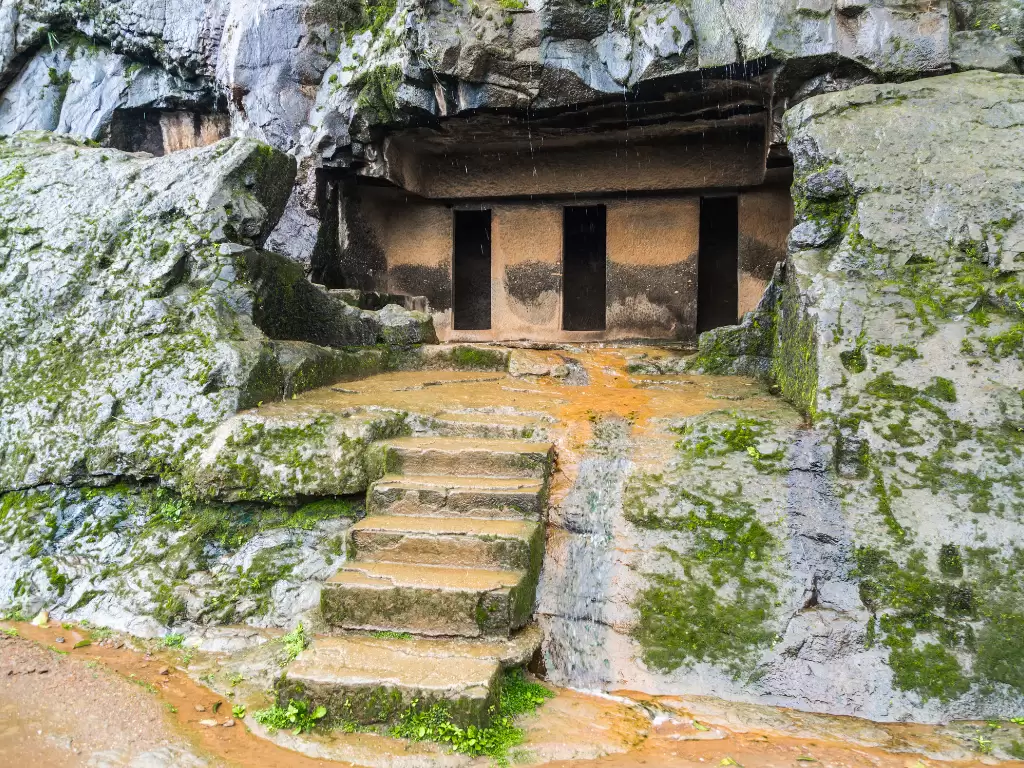 Ancient cave entrance with three doors at Bhaja Buddhist Caves during rainy mist, featuring stone steps mossy rocks, perfect heritage experience Pune caves tour package. 