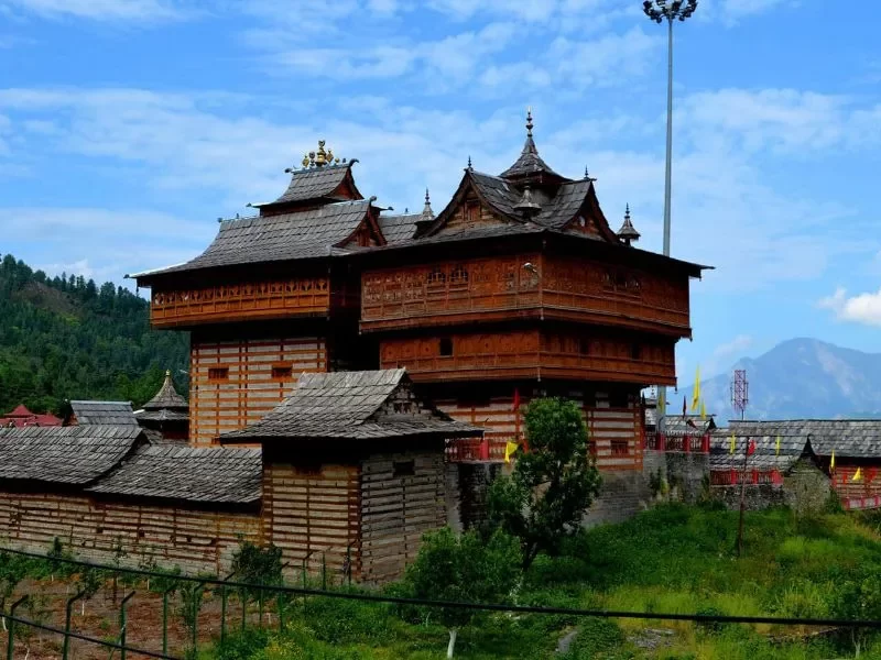 Shangchul Mahadev Temple pagoda at Shangarh Meadows during partly cloudy skies, featuring ornate wooden tower flags surrounding buildings pine mountains meadows, perfect spiritual adventure Himachal Pradesh tour package.