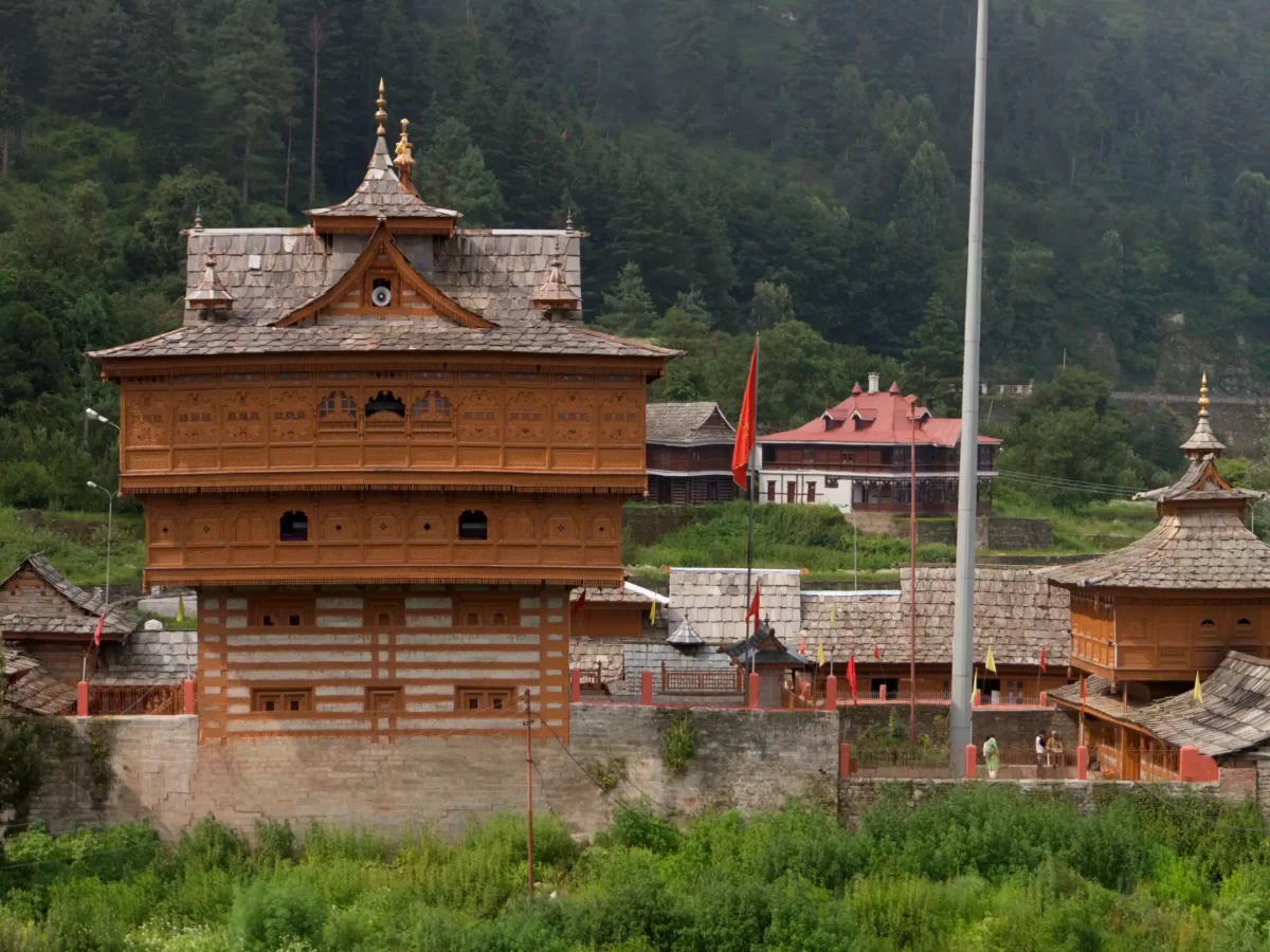 Shangchul Mahadev Temple complex at Shangarh Meadows during overcast weather, featuring multi-story wooden tower prayer flags green meadows pine forests mountains, perfect spiritual adventure Himachal Pradesh tour package.