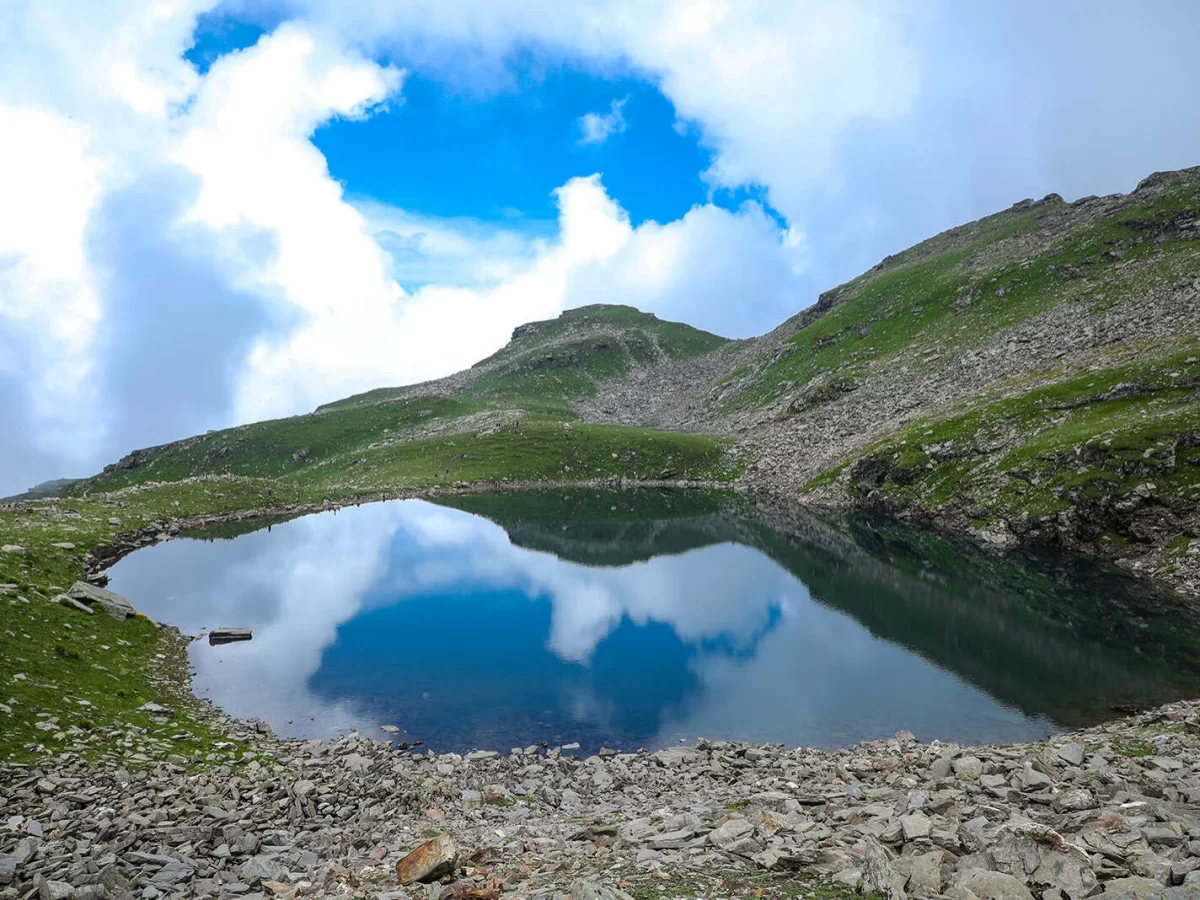 Bhrigu Lake at Manali Himachal Pradesh during partly cloudy skies, featuring mirror reflections green hills rocky shores, perfect trekking experience Himachal Pradesh tour package.