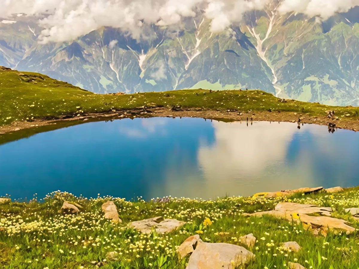 Bhrigu Lake at Manali Himachal Pradesh during cloudy skies, featuring turquoise water reflections snowy peaks alpine meadows wildflowers, perfect trekking experience Himachal Pradesh tour package.