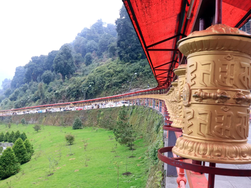 Prayer wheels corridor at Buddha Park of Ravangla during cloudy weather, featuring golden wheels and terraced green lawns, perfect spiritual Sikkim tour package.