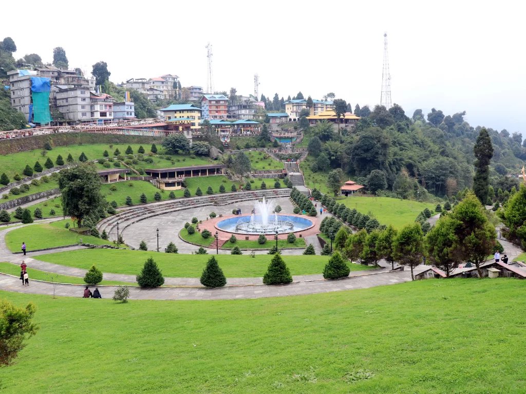 Terraced gardens and central fountain at Buddha Park of Ravangla during cloudy daylight, featuring green lawns, conifer rows and hillside town backdrop, perfect spiritual Sikkim tour package.