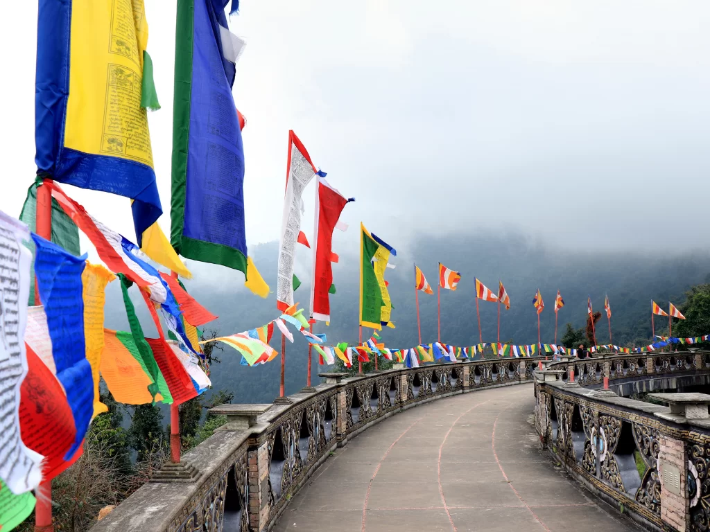 Prayer flag-lined pathway at Buddha Park of Ravangla during misty weather, featuring colorful flags and foggy mountain backdrop, perfect spiritual Sikkim tour package.