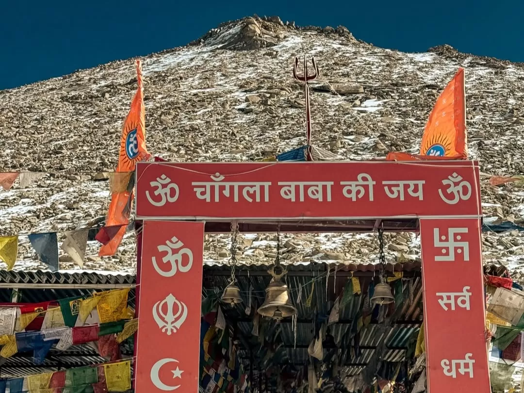Changla Pass shrine gate Chongla Baba Ki Jay during clear skies, featuring bells, prayer flags, saffron flags, perfect spiritual adventure Ladakh tour pack