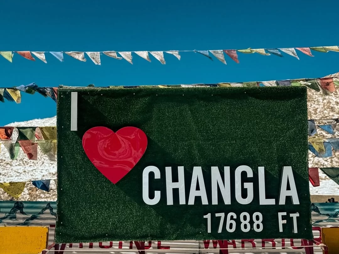 Changla Pass signboard at 17688 ft during sunny skies, featuring prayer flags, green turf backdrop, perfect high-altitude adventure Ladakh tour package.
