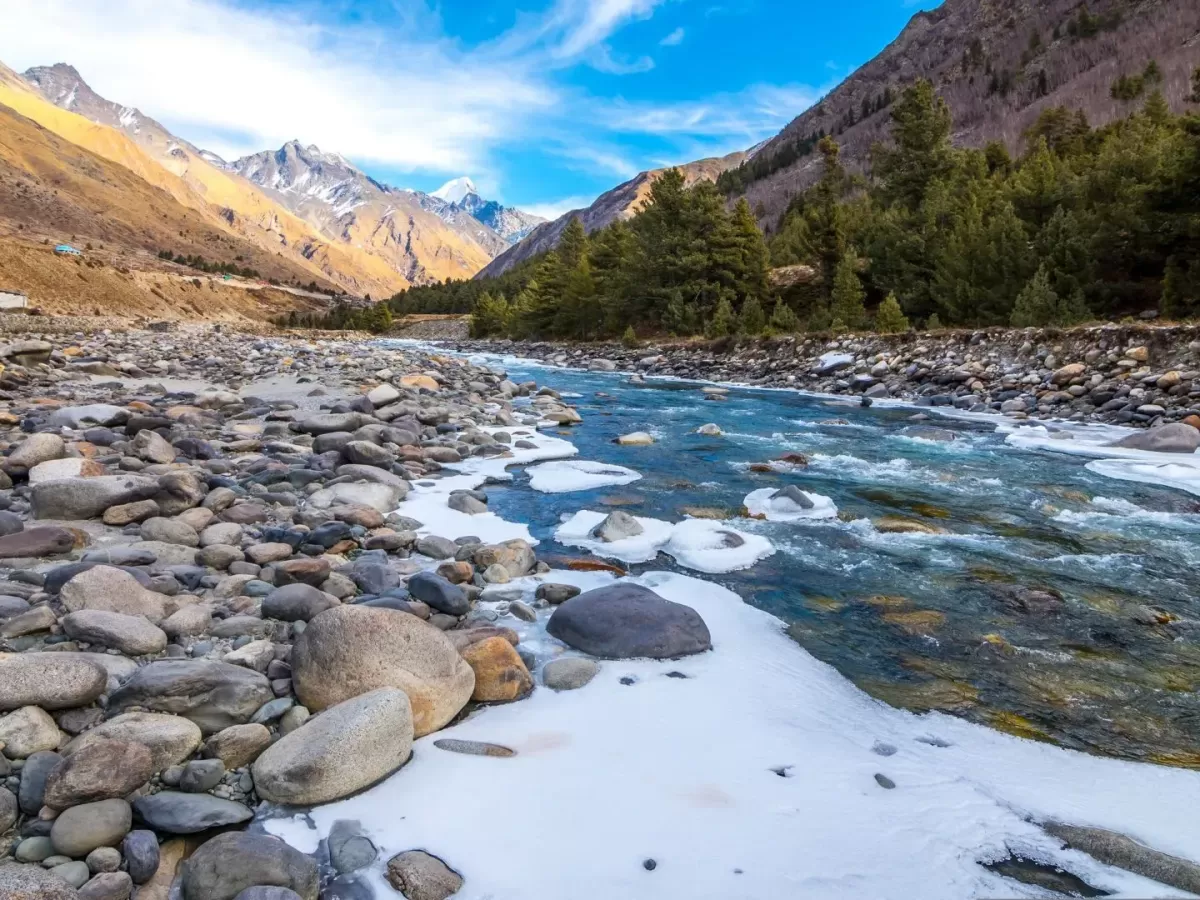 Baspa River Chitkul Village Kinnaur Himachal Pradesh during partly cloudy blue skies, featuring turquoise river snowy banks rocky riverbed pine forested snowy peaks valley backdrop, perfect cultural experience Himachal tour package.