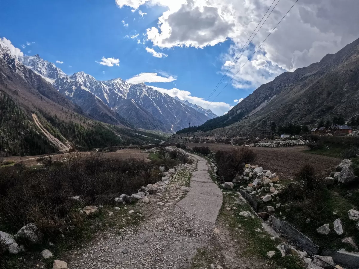 Chitkul Village Kinnaur Himachal Pradesh during partly cloudy blue skies, featuring stone pathway terraced fields snow-capped Himalayan peaks forested valley backdrop, perfect cultural experience Himachal tour package.