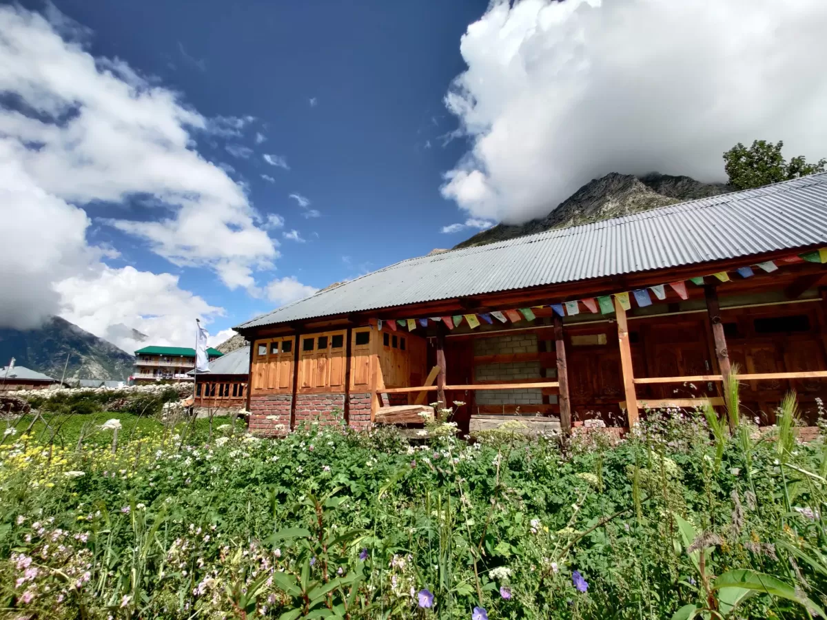 Chitkul Village Kinnaur Himachal Pradesh during partly cloudy blue skies, featuring wooden homestay prayer flags mountain backdrop wildflowers meadow foreground, perfect cultural experience Himachal tour package.