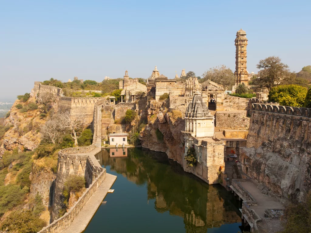 Vijay Stambh and cliffside temples at Chittorgarh Fort in Rajasthan during sunny day, featuring reservoir and stone walls, perfect Rajasthan tour package.