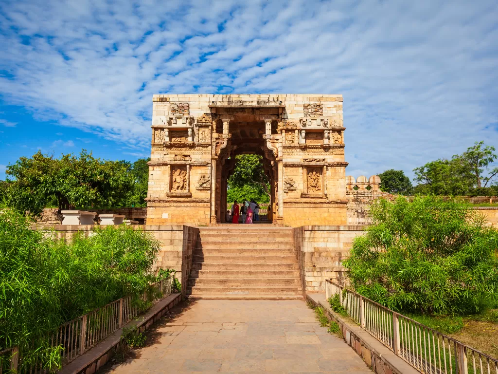 Ornate gateway at Chittorgarh Fort in Rajasthan under blue sky, featuring stone carvings and greenery, perfect Rajasthan